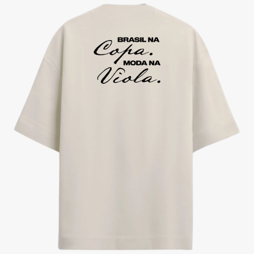 Camiseta com estampa frontal de brasão com um círculo e cinco estrelas acima, e estampa traseira de texto "Brasil na Copa. Moda na Viola." em estilo cursivo.