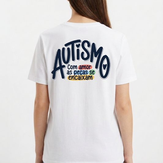 Camiseta com estampa frontal de mãos formando coração com peças de quebra-cabeça coloridas e estampa traseira com frase sobre autismo em diferentes cores.