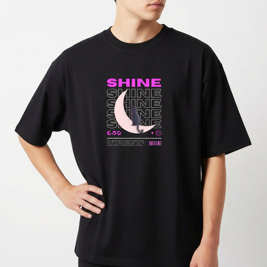 Camiseta com estampa frontal de texto "SHINE" repetido em camadas, sobreposto a uma meia-lua detalhada e pequena figura de um gato.