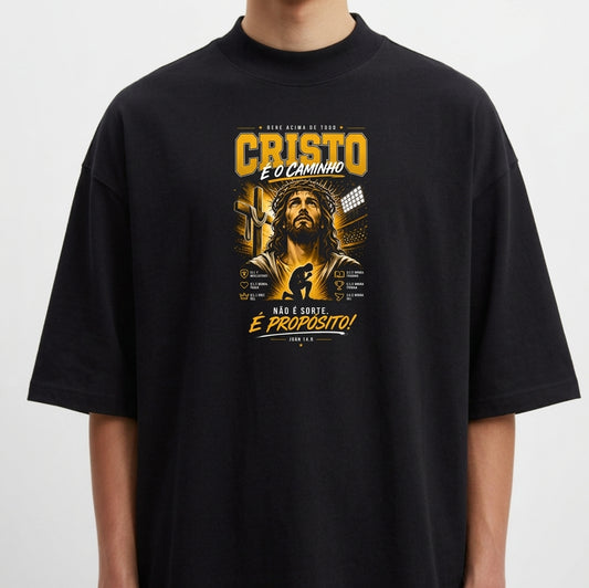 Camiseta com estampa frontal de uma figura religiosa cercada por textos e elementos gráficos detalhados, incluindo frases em destaque e simbologia variada.