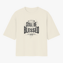 Camiseta com estampa frontal de texto 'Still Be Blessed' acompanhado de um halo, e estampa traseira de texto estilizado 'God is Good'.