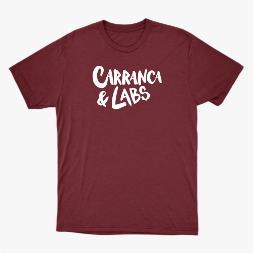 Camiseta com estampa frontal de letras estilizadas "Carranca & Labs" e estampa traseira de um desenho gráfico de figura caricata com expressão marcante.