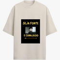 Camiseta com estampa frontal de personagem de cabelo curto em estilo desenho animado com mensagem inspiradora "SEJA FORTE E CORAJOSO" em letras destacadas.