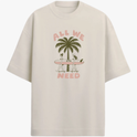 Camiseta com estampa frontal de duas pessoas carregando uma prancha de surfe diante de um coqueiro, acompanhada pela frase "ALL WE NEED".