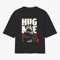 Camiseta com estampa frontal de tubarão antropomorfizado de braços cruzados, sobreposto a letras grandes "HUG ME" e fundo abstrato geométrico.