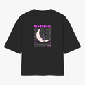 Camiseta com estampa frontal de texto "SHINE" repetido em camadas, sobreposto a uma meia-lua detalhada e pequena figura de um gato.