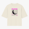 Camiseta com estampa frontal de texto "SHINE" repetido em camadas, sobreposto a uma meia-lua detalhada e pequena figura de um gato.