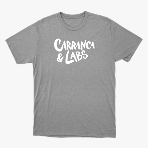 Camiseta com estampa frontal de letras estilizadas "Carranca & Labs" e estampa traseira de um desenho gráfico de figura caricata com expressão marcante.