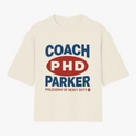 Camiseta com estampa frontal de texto em destaque centralizado, incluindo letras maiúsculas "COACH PHD PARKER" e frase complementar na parte inferior.