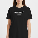 Camiseta com estampa frontal de texto grande "OBSESSED" seguido por linhas menores que incluem mensagens inspiradoras e dois logotipos pequenos abaixo.