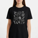 Camiseta com estampa frontal de desenhos sorridentes de criaturas vegetais estilizadas, cercadas por delicados detalhes de flores e folhagens diversas.