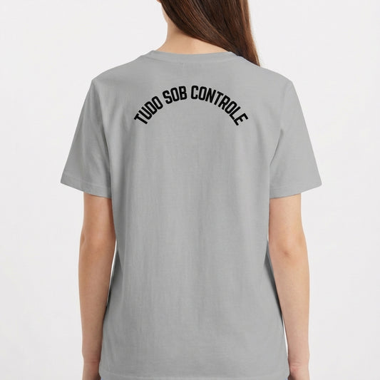 Camiseta com estampa frontal de gato preto segurando um copo, envolto em formas abstratas, e estampa traseira com texto em arco "TUDO SOB CONTROLE".
