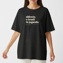 Camiseta com estampa frontal de texto "silêncio. o brasil tá jogando." com assinatura "FALAR, NÃO MURMURAR." e estampa traseira de "VERDELO" no topo.