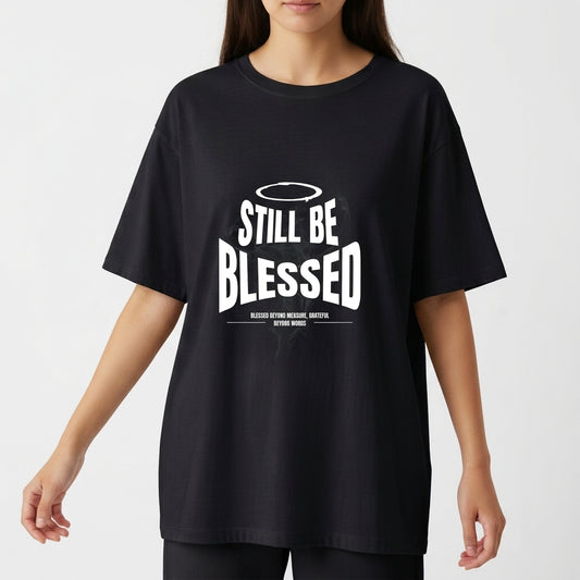 Camiseta com estampa frontal de texto 'Still Be Blessed' acompanhado de um halo, e estampa traseira de texto estilizado 'God is Good'.