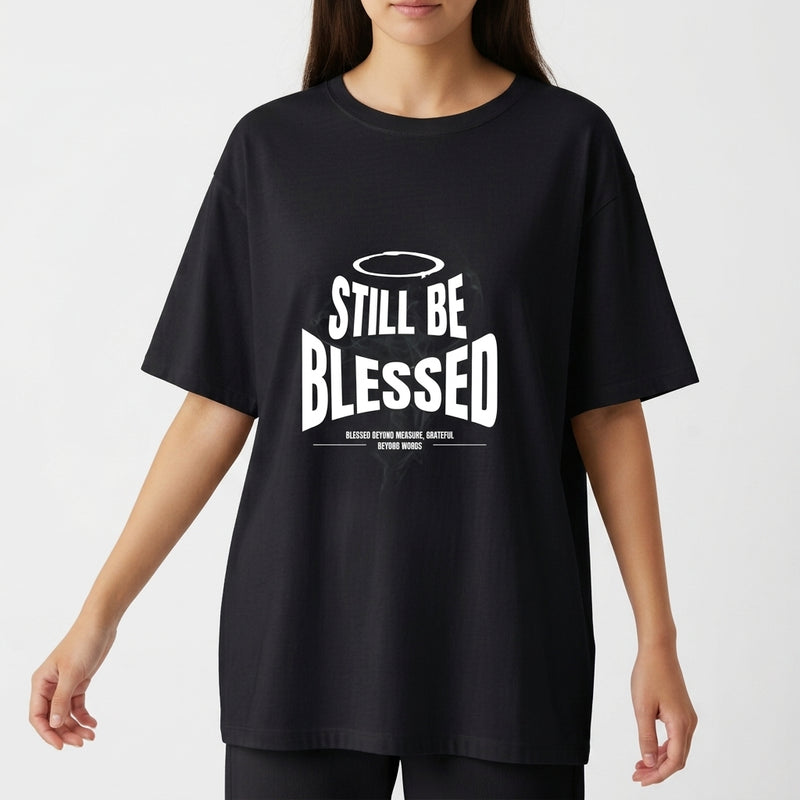 Camiseta com estampa frontal de texto 'Still Be Blessed' acompanhado de um halo, e estampa traseira de texto estilizado 'God is Good'.