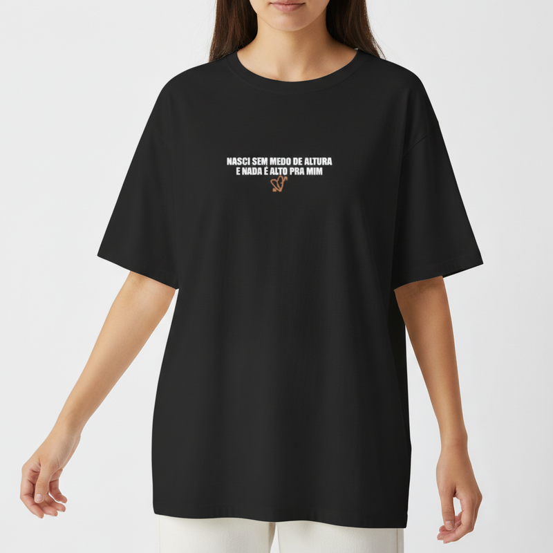 Camiseta com estampa frontal de texto "Nasci sem medo de altura e nada é alto pra mim" com detalhes gráficos e estampa traseira de mãos tatuadas com anéis.
