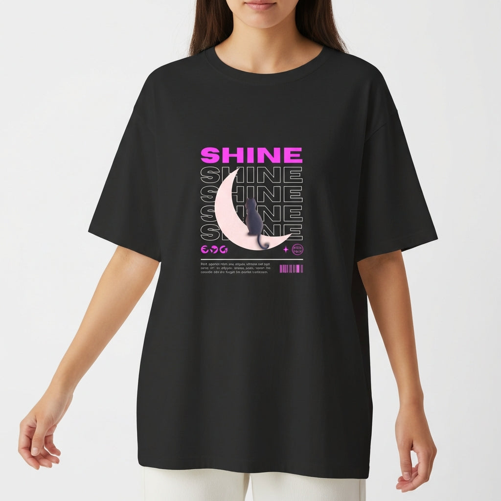 Camiseta com estampa frontal de texto "SHINE" repetido em camadas, sobreposto a uma meia-lua detalhada e pequena figura de um gato.