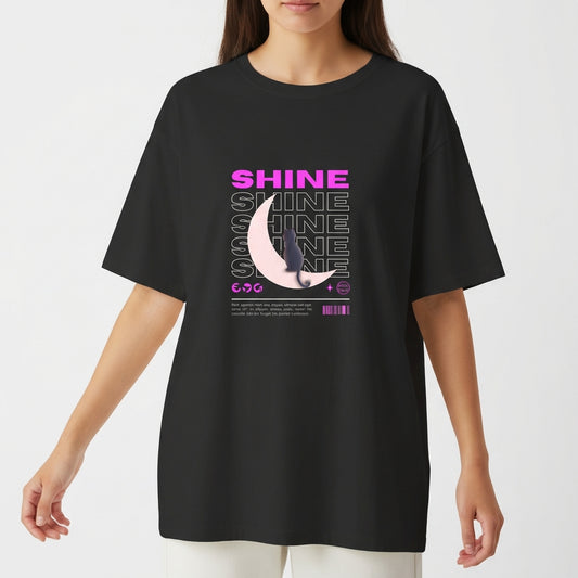 Camiseta com estampa frontal de texto "SHINE" repetido em camadas, sobreposto a uma meia-lua detalhada e pequena figura de um gato.