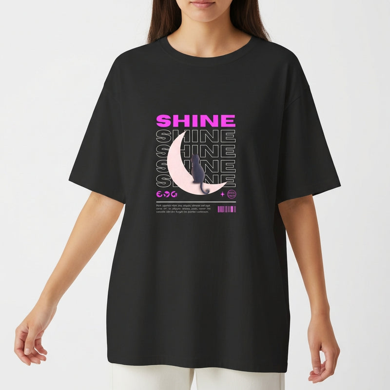 Camiseta com estampa frontal de texto "SHINE" repetido em camadas, sobreposto a uma meia-lua detalhada e pequena figura de um gato.