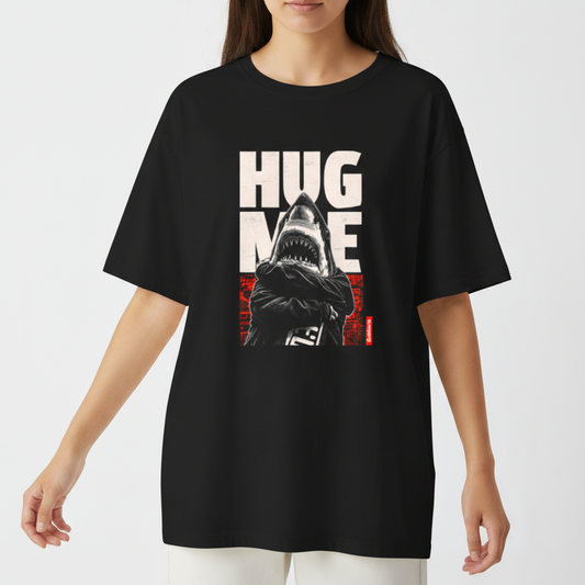 Camiseta com estampa frontal de tubarão antropomorfizado de braços cruzados, sobreposto a letras grandes "HUG ME" e fundo abstrato geométrico.