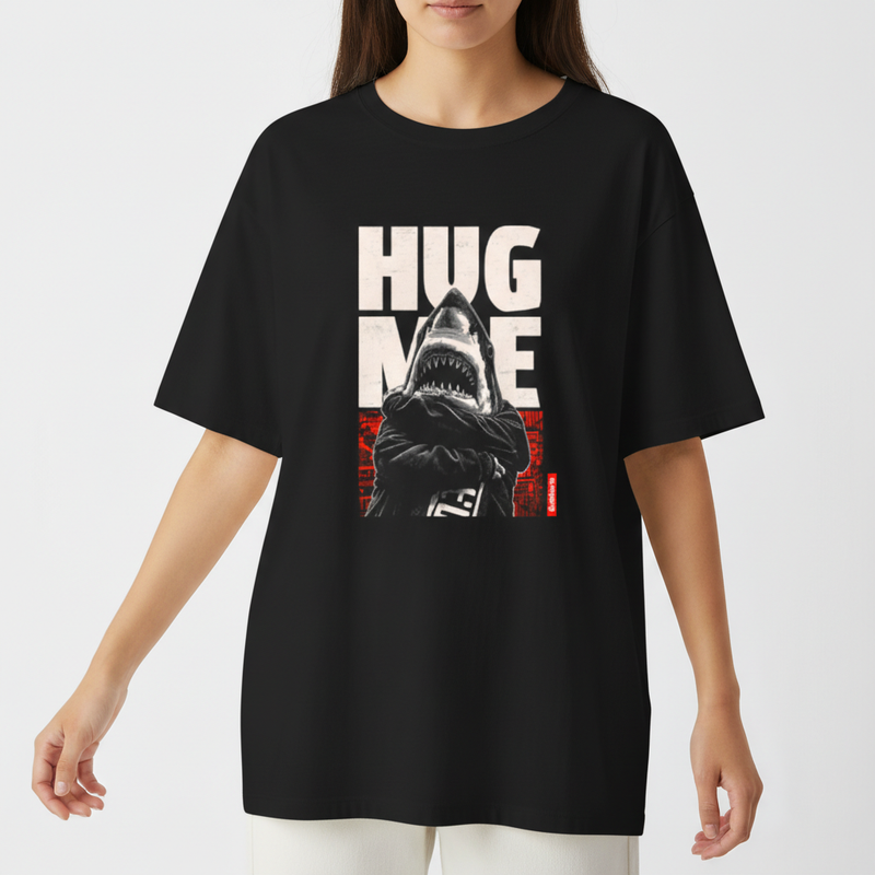 Camiseta com estampa frontal de tubarão antropomorfizado de braços cruzados, sobreposto a letras grandes "HUG ME" e fundo abstrato geométrico.