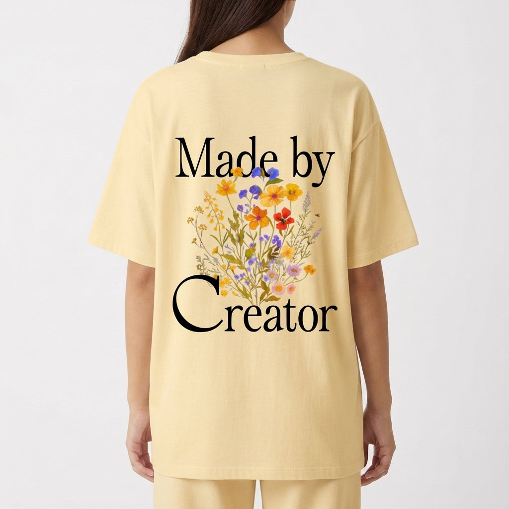 Camiseta com estampa frontal de "Made by Creator" em letras estilizadas e estampa traseira de "Made by Creator" acompanhada por ilustração de flores coloridas.
