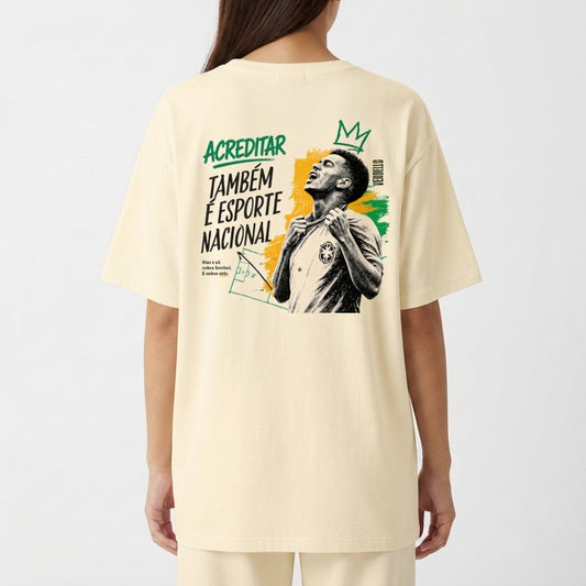 Camiseta com estampa frontal de pequena coroa e "ACREDITAR" estilizado, e estampa traseira de jogador com coroa, cores vibrantes e mensagem inspiradora.