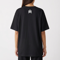 Camiseta com estampa frontal de rosto sorridente decorado com folhas e texto "Elevate Your Frequency" emoldurado por elementos naturais e estampa traseira de símbolo geométrico simples próximo à gola.