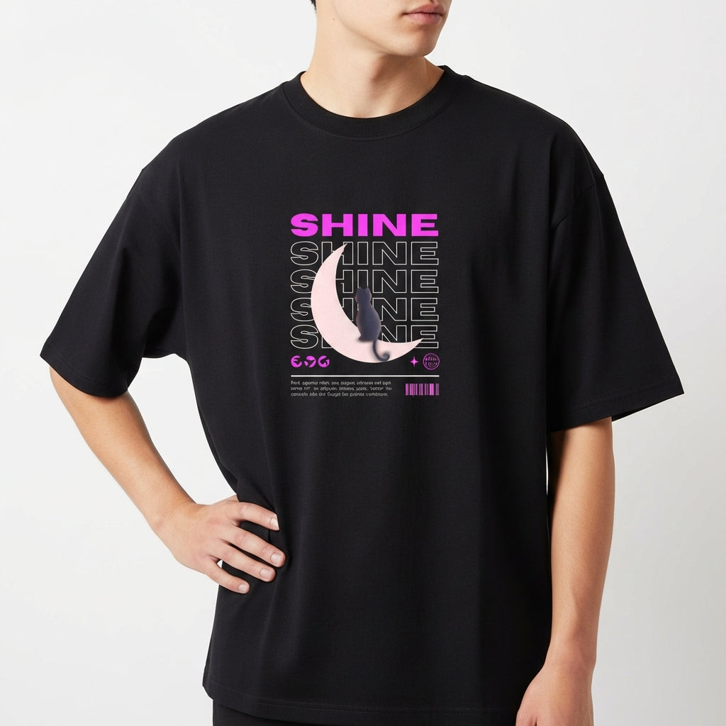 Camiseta com estampa frontal de texto "SHINE" repetido em camadas, sobreposto a uma meia-lua detalhada e pequena figura de um gato.
