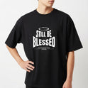 Camiseta com estampa frontal de texto 'Still Be Blessed' acompanhado de um halo, e estampa traseira de texto estilizado 'God is Good'.