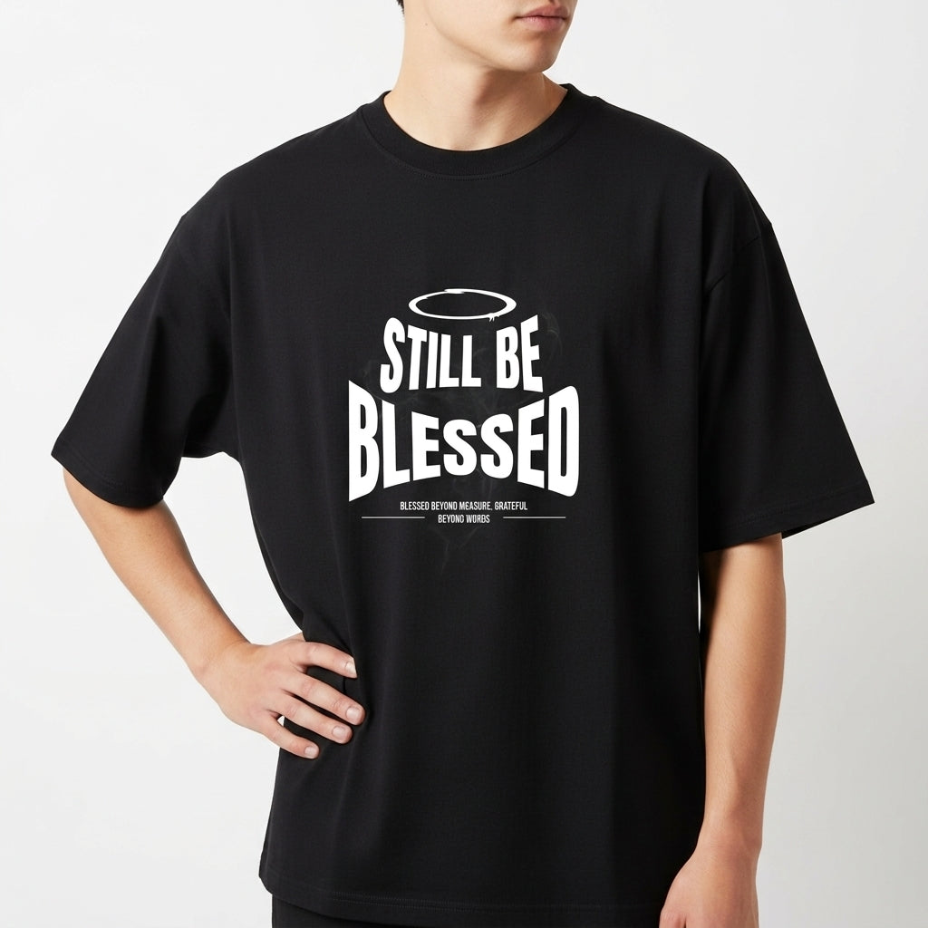Camiseta com estampa frontal de texto 'Still Be Blessed' acompanhado de um halo, e estampa traseira de texto estilizado 'God is Good'.