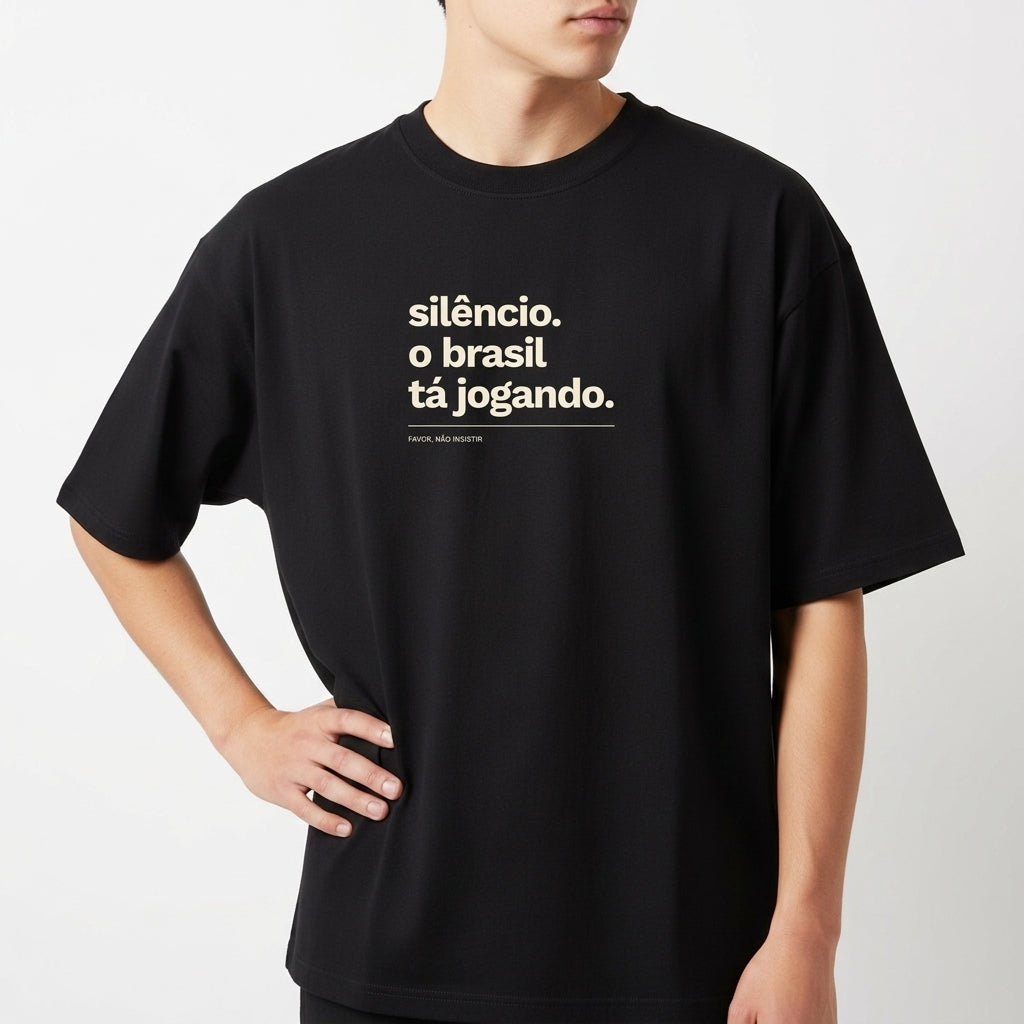 Camiseta com estampa frontal de texto "silêncio. o brasil tá jogando." com assinatura "FALAR, NÃO MURMURAR." e estampa traseira de "VERDELO" no topo.