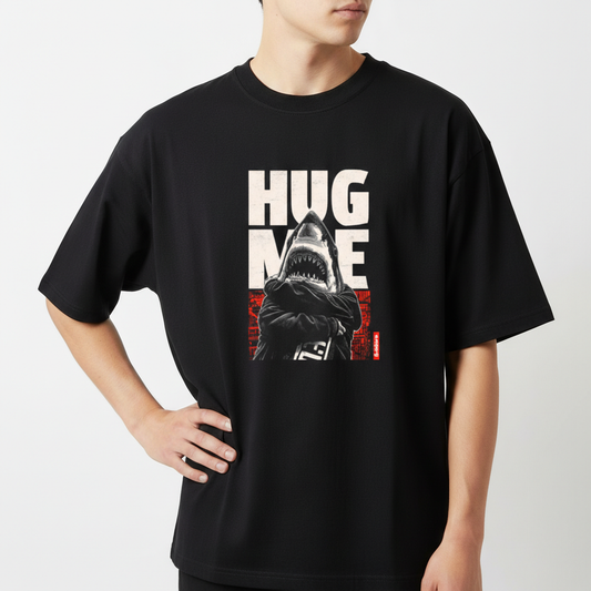 Camiseta com estampa frontal de tubarão antropomorfizado de braços cruzados, sobreposto a letras grandes "HUG ME" e fundo abstrato geométrico.