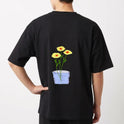 Camiseta com estampa frontal de texto em letras simples "Il raccolto è inevitabile." e estampa traseira de vaso azul com três flores amarelas e caule verde.