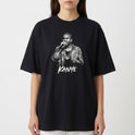Camiseta com estampa frontal de um cantor segurando um microfone, vestindo jaqueta e colares, acompanhada pelo nome "Kanye" estilizado abaixo.