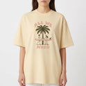 Camiseta com estampa frontal de duas pessoas carregando uma prancha de surfe diante de um coqueiro, acompanhada pela frase "ALL WE NEED".