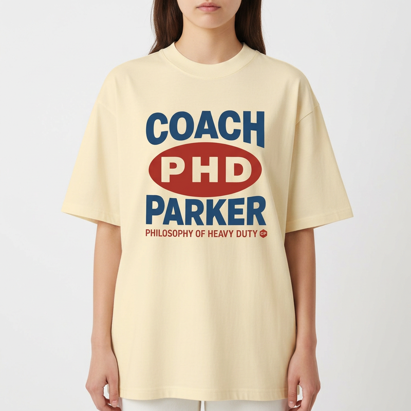 Camiseta com estampa frontal de texto estilizado com palavras "COACH PHD PARKER" em destaque, acompanhada de "PHILOSOPHY OF HEAVY DUTY" em menor tamanho.