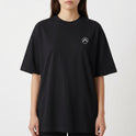 Camiseta com estampa frontal de símbolo minimalista circular com seta apontando para cima e estampa traseira de paisagem tropical com palmeiras e pôr do sol refletido no mar.