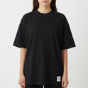 Camiseta com estampa frontal de logotipo minimalista e estampa traseira de prancha de surf encostada em parede desgastada com palmeira e fundo geométrico.