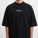 Camiseta com estampa frontal de logotipo estilizado com ornamentos e estampa traseira de figura esquelética sentada em um trono com detalhes rústicos.