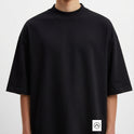 Camiseta com estampa frontal de logotipo minimalista e estampa traseira de prancha de surf encostada em parede desgastada com palmeira e fundo geométrico.
