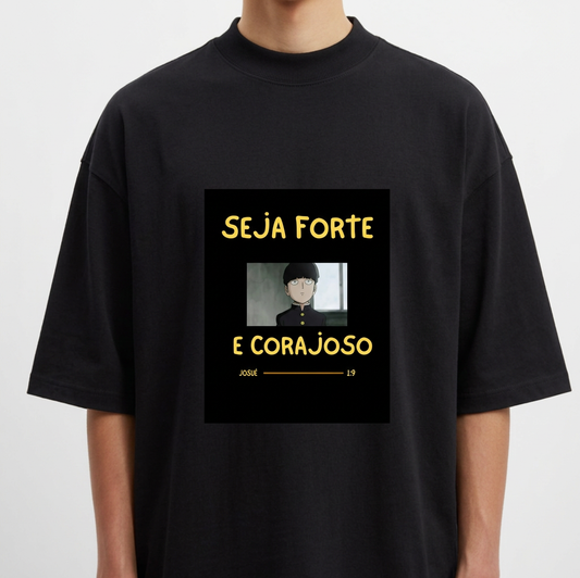 Camiseta com estampa frontal de personagem de cabelo curto em estilo desenho animado com mensagem inspiradora "SEJA FORTE E CORAJOSO" em letras destacadas.