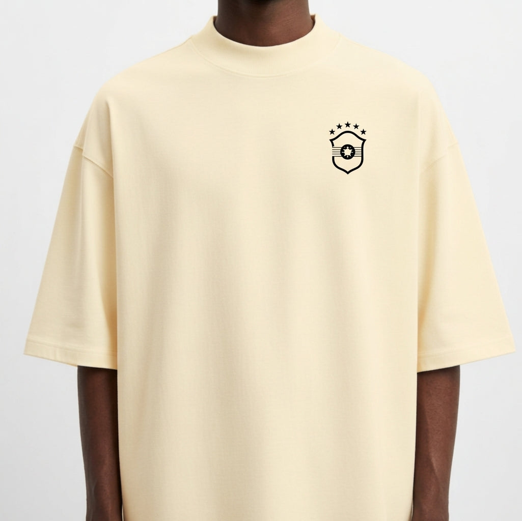 Camiseta com estampa frontal de brasão com um círculo e cinco estrelas acima, e estampa traseira de texto "Brasil na Copa. Moda na Viola." em estilo cursivo.