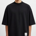 Camiseta com estampa frontal de pequeno logotipo minimalista e estampa traseira de folhas tropicais em diversas cores e círculo texturizado.