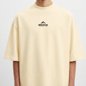 Camiseta com estampa frontal de montanha e texto sob ela, e estampa traseira detalhada de figura mitológica central cercada por caveiras e chamas.