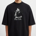 Camiseta com estampa frontal de um artista cantando no microfone, vestindo boné e camisa preta, destacando tatuagens nos braços e autógrafo abaixo.