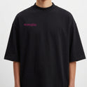 Camiseta com estampa frontal de inscrição estilizada e estampa traseira de urso sorridente com olhos em forma de "X" sobre fundo de respingos vibrantes.