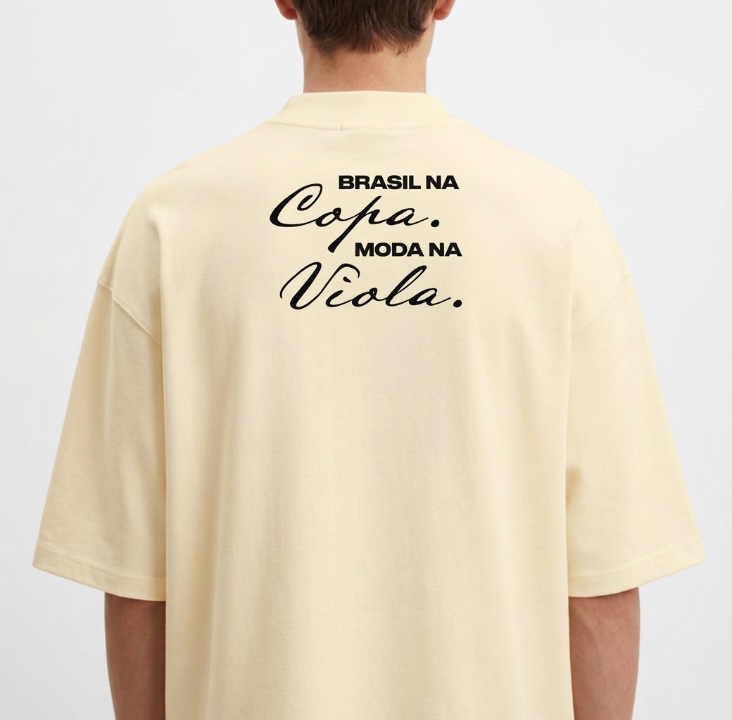 Camiseta com estampa frontal de brasão com um círculo e cinco estrelas acima, e estampa traseira de texto "Brasil na Copa. Moda na Viola." em estilo cursivo.