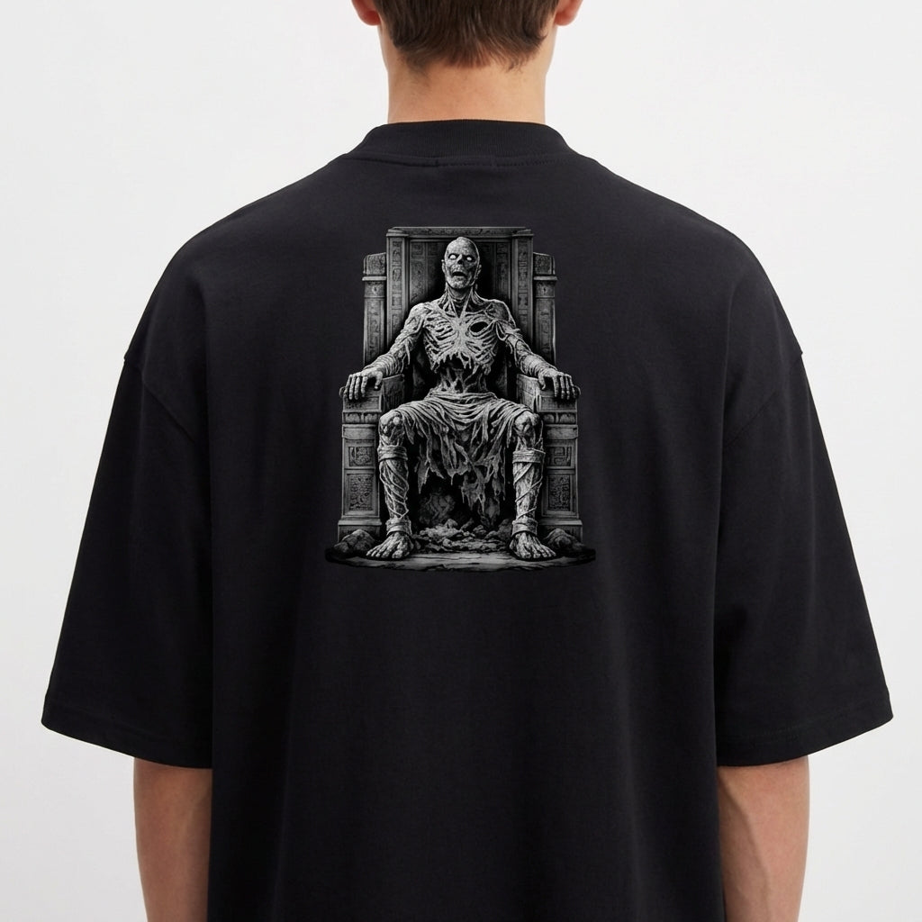 Camiseta com estampa frontal de logotipo estilizado com ornamentos e estampa traseira de figura esquelética sentada em um trono com detalhes rústicos.