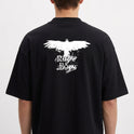 Camiseta com estampa frontal de caveira pequena e estampa traseira de pássaro em voo com texto estilizado por baixo, criando um contraste marcante.